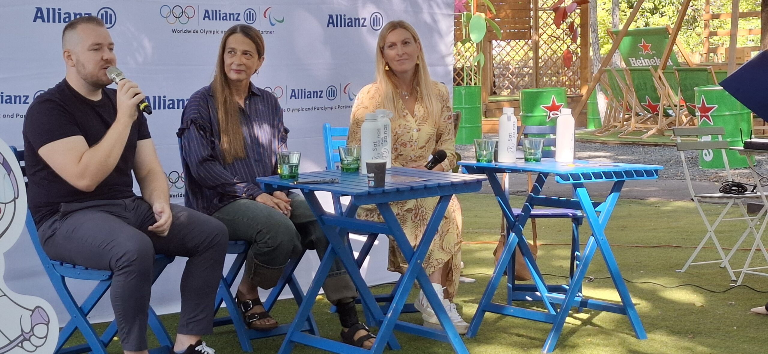 Fotografija prikazuje troje ljudi od kojih jedan drži mikrofon, a dvije djevojke sjede pokraj njega. Iza leđa nazi se plakat na kojem piše 'Allianz'.