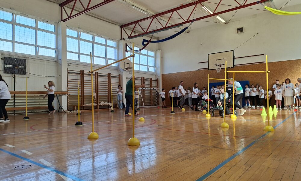 Fotografija prikazuje živahnu atmosferu u školskoj sportskoj dvorani, ispunjenoj djecom i odraslima koji sudjeluju u organiziranoj tjelesnoj aktivnosti. Prostor je prostran, s sjajnim drvenim podom obilježenim crvenim i plavim linijama za sportske terene. Visoki prozori propuštaju obilje prirodnog svjetla, dok crvene metalne grede na stropu daju industrijski, ali topao dojam. U središtu dvorane postavljeni su žuti stupići s okruglim bazama, raspoređeni u uzorku koji podsjeća na poligon za vježbanje ili igru. Uz njih se nalaze žuti čunjevi u ravnoj liniji, a na zidu je košarkaški obruč. Djeca u bijelim majicama stoje uz zid, dok su druga aktivno uključena u igru. Posebno dirljiv trenutak prikazuje odraslu osobu koja pomaže djetetu u invalidskim kolicima da prođe kroz poligon — snažan prikaz inkluzije i zajedništva.