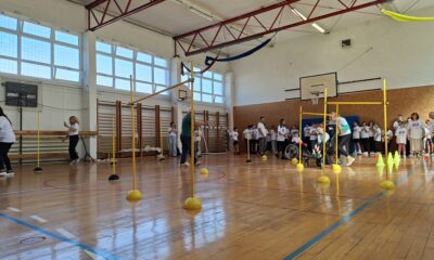 Fotografija prikazuje živahnu atmosferu u školskoj sportskoj dvorani, ispunjenoj djecom i odraslima koji sudjeluju u organiziranoj tjelesnoj aktivnosti. Prostor je prostran, s sjajnim drvenim podom obilježenim crvenim i plavim linijama za sportske terene. Visoki prozori propuštaju obilje prirodnog svjetla, dok crvene metalne grede na stropu daju industrijski, ali topao dojam. U središtu dvorane postavljeni su žuti stupići s okruglim bazama, raspoređeni u uzorku koji podsjeća na poligon za vježbanje ili igru. Uz njih se nalaze žuti čunjevi u ravnoj liniji, a na zidu je košarkaški obruč. Djeca u bijelim majicama stoje uz zid, dok su druga aktivno uključena u igru. Posebno dirljiv trenutak prikazuje odraslu osobu koja pomaže djetetu u invalidskim kolicima da prođe kroz poligon — snažan prikaz inkluzije i zajedništva.