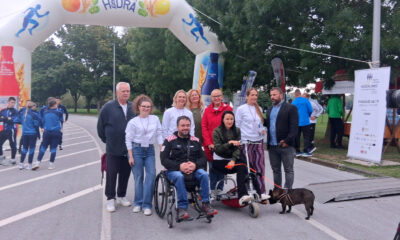 Skupina ljudi pozira ispod velikog napuhanog luka s logom Hidra tijekom akcije „Hodajmo za one koji ne mogu“. U prvom planu je muškarac u invalidskim kolicima i žena na triciklu s psom, okruženi volonterima i sudionicima događaja.