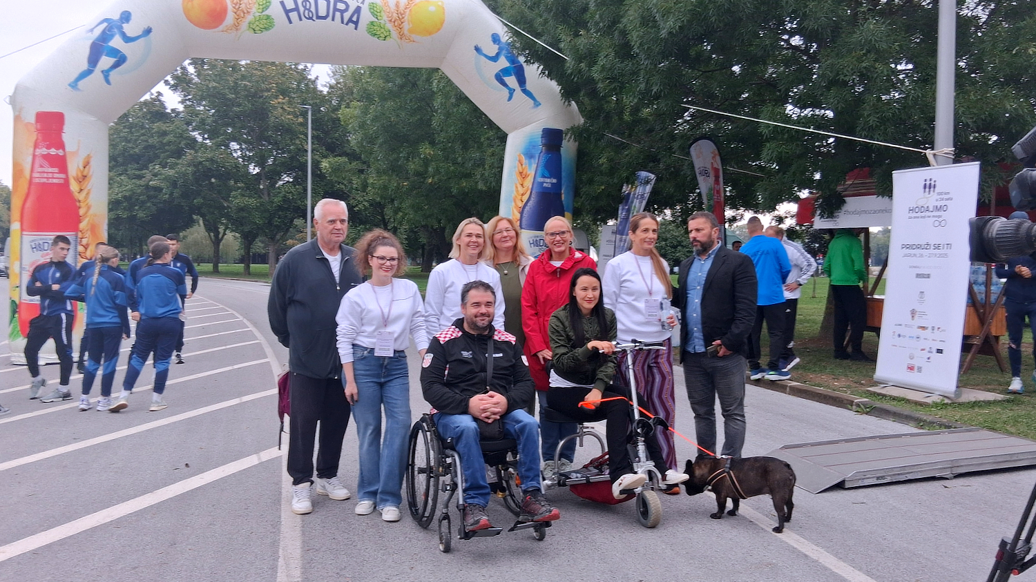 Skupina ljudi pozira ispod velikog napuhanog luka s logom Hidra tijekom akcije „Hodajmo za one koji ne mogu“. U prvom planu je muškarac u invalidskim kolicima i žena na triciklu s psom, okruženi volonterima i sudionicima događaja.