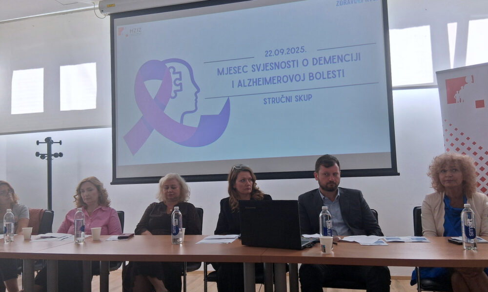 Na slici je stručni skup povodom Mjeseca svjesnosti o demenciji i Alzheimerovoj bolesti, održan 22. rujna 2025. godine. Ispred projekcijskog platna na kojem je prikazan ljubičasti logo u obliku vrpce i profila ljudske glave sjedi šestero sudionika, tri žene s lijeve strane, dvije žene i jedan muškarac s desne strane. Na stolu ispred njih nalaze se boce vode, papiri i prijenosno računalo.