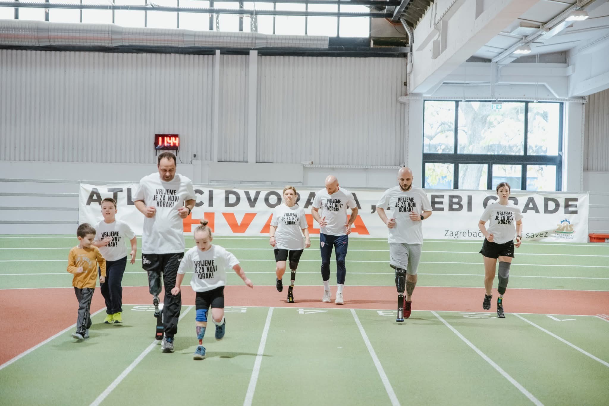 Fotografija prikazuje nekoliko osoba s protezama kako trče po atletskoj stazi u dvorani.