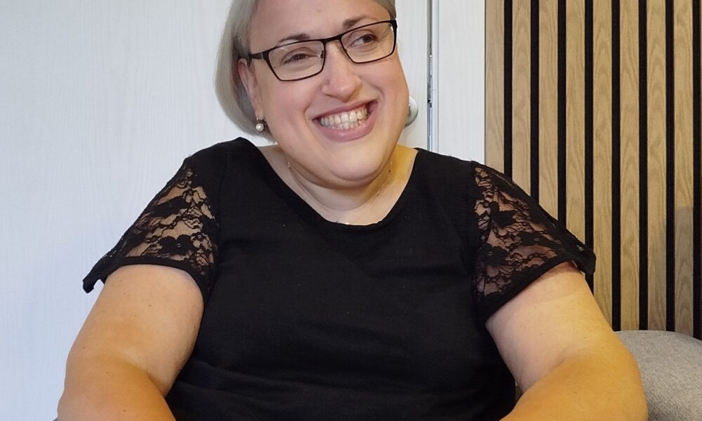 Na fotografiji se nalazi osoba koja sjedi u invalidskim kolicima i veselo se smiješi prema kameri. Ima kratku sijedu kosu, nosi naočale i odjevena je u crnu majicu s čipkastim rukavima. Na lijevom zapešću nosi narukvicu s privjescima. U pozadini se vidi bijela vrata i zid obložen drvenim panelima. Atmosfera fotografije djeluje toplo i prijateljski.