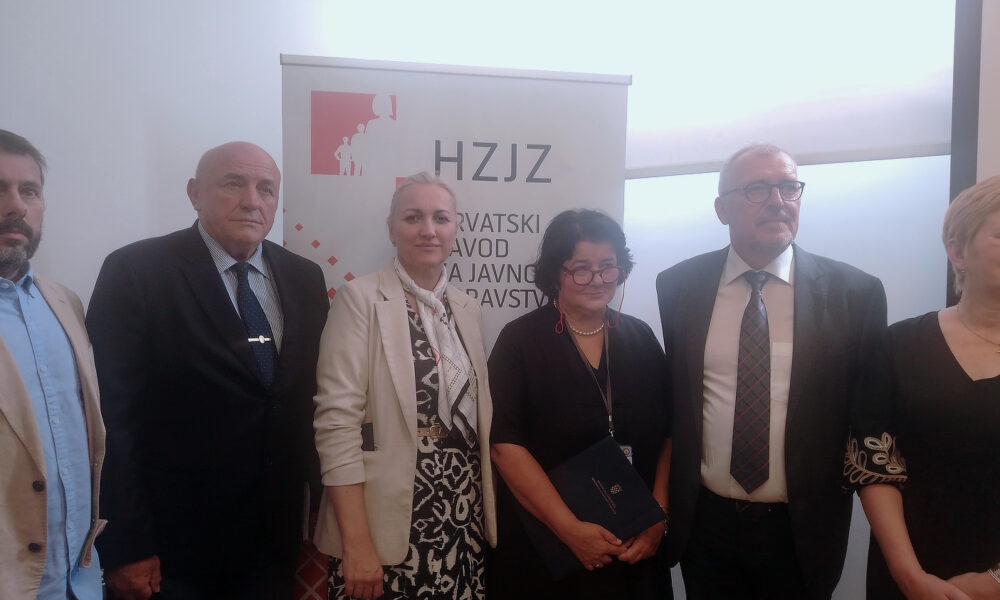 Skupina muškaraca i žena u poslovnoj odjeći stoji ispred roll-up plakata s logotipom i natpisom 'HZJZ – Hrvatski zavod za javno zdravstvo'. Nalaze se u zatvorenom prostoru, pozirajući za zajedničku fotografiju.