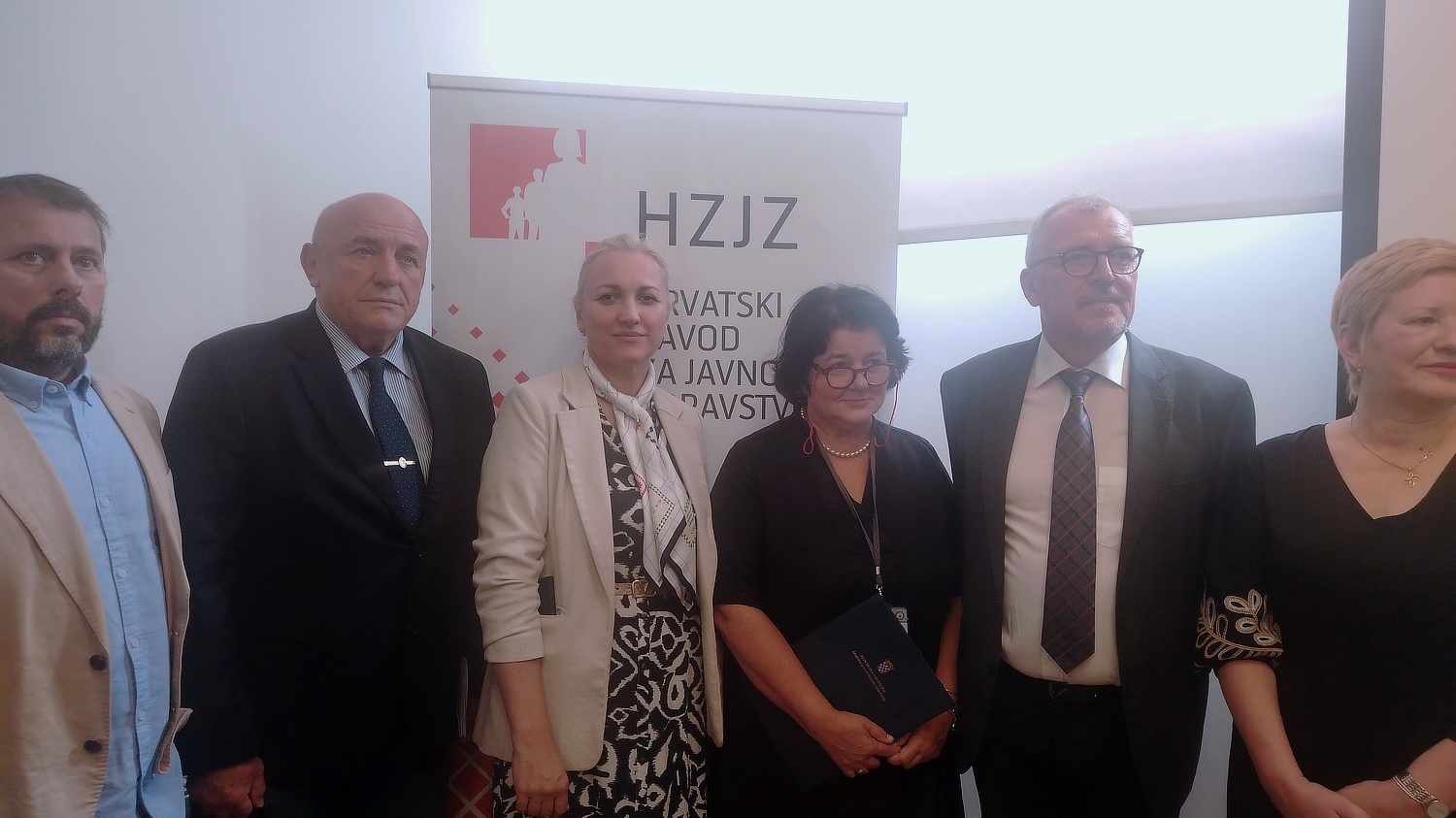 Skupina muškaraca i žena u poslovnoj odjeći stoji ispred roll-up plakata s logotipom i natpisom 'HZJZ – Hrvatski zavod za javno zdravstvo'. Nalaze se u zatvorenom prostoru, pozirajući za zajedničku fotografiju.