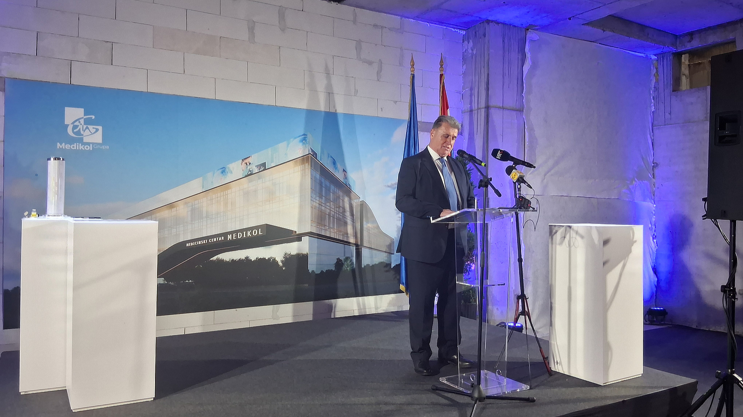Muškarac u odijelu stoji za govornicom i drži govor na svečanosti. Iza njega se nalazi velika slika buduće zgrade s natpisom 'Medicinski centar Medikol'. U prostoru se vide hrvatska i europska zastava te tehnička oprema za ozvučenje i snimanje.