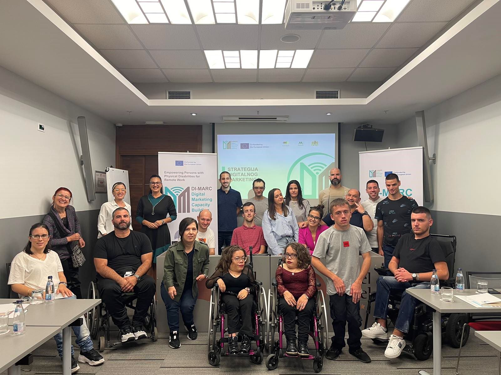 Na slici se nalazi skupina od dvadeset ljudi okupljenih u konferencijskoj ili seminarijskoj prostoriji. Neki sjede, neki stoje, a nekoliko osoba koristi invalidska kolica. Atmosfera je profesionalna i inkluzivna, što sugerira da se radi o radionici, seminaru ili treningu usmjerenom na pristupačnost, liderstvo ili društveni angažman. U pozadini se vide plakati i projekcijsko platno s logotipima i tekstom.
