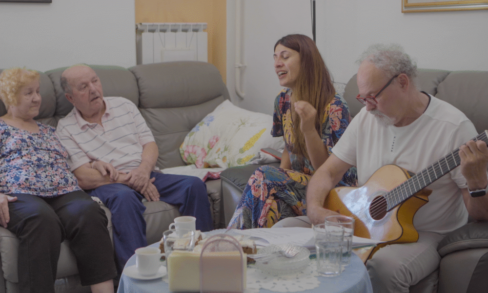 Na slici su četiri odrasle osobe koje sjede u dnevnoj sobi, u toplom i intimnom okruženju koje odiše obiteljskom atmosferom. Na lijevoj strani sjede stariji muškarac i žena na sivoj garnituri. Žena ima kratku kovrčavu kosu, nosi bluzu s uzorkom i crne hlače. Muškarac je odjeven u prugastu polo majicu i tamne hlače. Do njih sjedi mlađa žena u cvjetnoj haljini, koja izgleda kao da pjeva ili strastveno govori – njezin izraz lica i držanje prenose snažnu emociju. Na desnoj strani nalazi se muškarac sijede kose i s naočalama, koji svira klasičnu gitaru. Odjeven je u bijelu košulju i svijetle hlače. Ambijent: Ispred njih je stolić s čašom vode, tanjurom s hranom, držačem za salvete i ukrasnim miljeom. U pozadini se vidi radijator i jastuk s cvjetnim uzorkom na kauču. Sve skupa sugerira neformalno, ugodno druženje – možda obiteljsko okupljanje ili večer s prijateljima uz glazbu i razgovor.