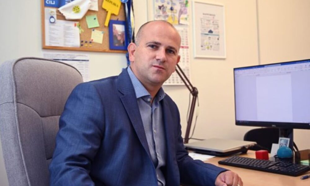 Na fotografiji se nalazi osoba u uredskom okruženju, sjedi za radnim stolom i gleda ravno u kameru. Odjevena je u plavi poslovni odijelo, što ostavlja profesionalan dojam. Na stolu se nalaze: • monitor s praznim dokumentom na ekranu • tipkovnica i miš • crvena antistres loptica • plavi predmet (možda držač ili ukras) • uokvirena fotografija – moguće osobna uspomena.