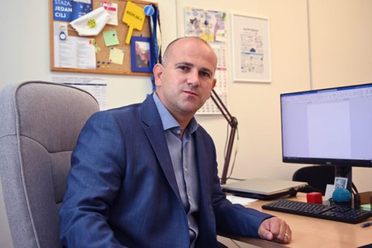 Na fotografiji se nalazi osoba u uredskom okruženju, sjedi za radnim stolom i gleda ravno u kameru. Odjevena je u plavi poslovni odijelo, što ostavlja profesionalan dojam. Na stolu se nalaze: • monitor s praznim dokumentom na ekranu • tipkovnica i miš • crvena antistres loptica • plavi predmet (možda držač ili ukras) • uokvirena fotografija – moguće osobna uspomena.