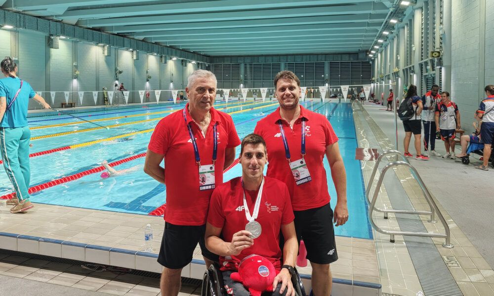 Na slici je hrvatski paraolimpijski plivač u kolicima koji ponosno pokazuje srebrnu medalju. Uz njega stoje dva trenera u crvenim majicama. Fotografija je snimljena kraj unutarnjeg bazena, u pozadini se vidi nekoliko plivača u vodi i drugih sportaša uz rub bazena.
