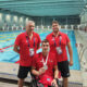 Na slici je hrvatski paraolimpijski plivač u kolicima koji ponosno pokazuje srebrnu medalju. Uz njega stoje dva trenera u crvenim majicama. Fotografija je snimljena kraj unutarnjeg bazena, u pozadini se vidi nekoliko plivača u vodi i drugih sportaša uz rub bazena.