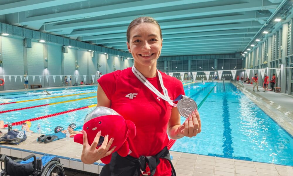 Fotografija prikazuje djevojku koja ima srebrnu medalju oko vrata, a u ruci drži neku igračku, dok se iza nje nalzi bazen.
