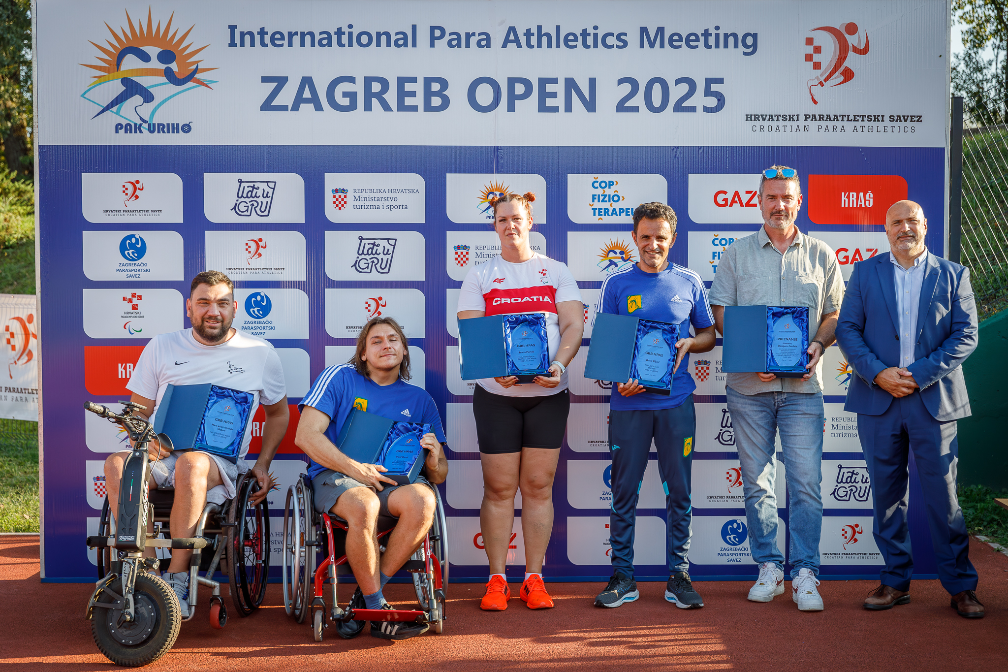 Fotografija prikazuje nekoliko ljudi koji su dobili priznanja. Neki su u invalidskim kolicima neki stoje. Iznad sportaša piše 'Internatinal para atletics Metting Zagreb 2025'.