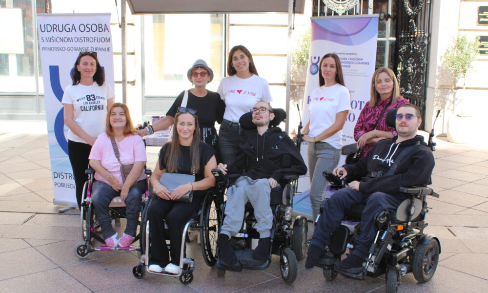 Na fotografiji je skupina članova i suradnika Udruge osoba s mišićnom distrofijom Primorsko-goranske županije. Nekoliko osoba sjedi u invalidskim kolicima, dok ostali stoje iza njih, svi nasmijani i vedri. U pozadini se nalaze promotivni roll-up plakati Udruge s logotipom i nazivom, a jasno se vidi da je riječ o javnom događaju održanom na otvorenom, najvjerojatnije na riječkom Korzu. Cijela scena odiše zajedništvom, pozitivnom energijom i naglaskom na inkluziju i podršku osobama s invaliditetom.