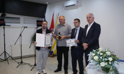 Na slici su četiri muškarca koji stoje u zatvorenom prostoru, najvjerojatnije na svečanosti dodjele priznanja ili nagrada. Dvojica drže certifikate, a jedan od njih i trofej. Prvi muškarac s lijeva drži veliki certifikat ili plaketu s tekstom i grafikom. Drugi muškarac drži trofej i certifikat s natpisom "SUBFIRA". Treći muškarac, odjeven u odijelo, također drži certifikat s istim natpisom. Četvrti muškarac, krajnje desno, stoji s rukama sklopljenim ispred sebe i ne drži ništa. U pozadini se vidi stol s cvjetnim aranžmanom, stalak za mikrofon, zvučnici i zastave, što dodatno naglašava formalnost događaja. Prostorija ima bijele zidove, pločice na podu, klima uređaje i prozore.