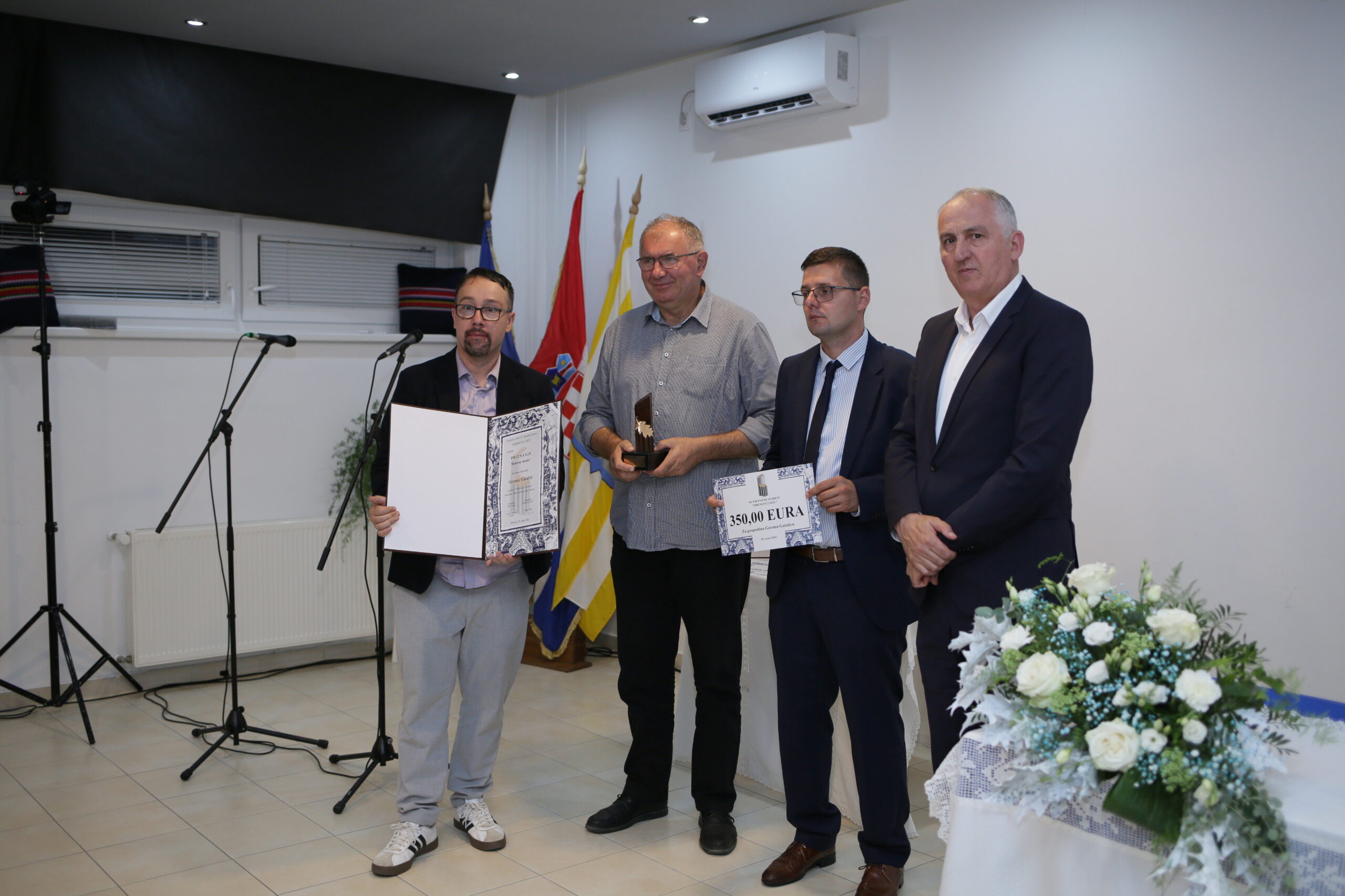 Na slici su četiri muškarca koji stoje u zatvorenom prostoru, najvjerojatnije na svečanosti dodjele priznanja ili nagrada. Dvojica drže certifikate, a jedan od njih i trofej. Prvi muškarac s lijeva drži veliki certifikat ili plaketu s tekstom i grafikom. Drugi muškarac drži trofej i certifikat s natpisom "SUBFIRA". Treći muškarac, odjeven u odijelo, također drži certifikat s istim natpisom. Četvrti muškarac, krajnje desno, stoji s rukama sklopljenim ispred sebe i ne drži ništa. U pozadini se vidi stol s cvjetnim aranžmanom, stalak za mikrofon, zvučnici i zastave, što dodatno naglašava formalnost događaja. Prostorija ima bijele zidove, pločice na podu, klima uređaje i prozore.