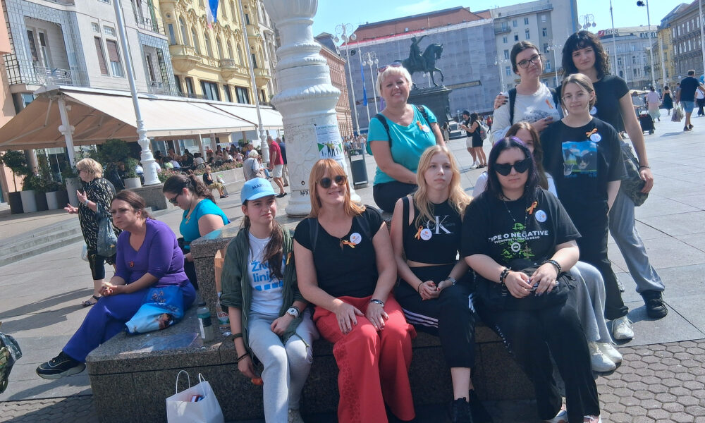 Grupa žena i djevojaka sjedi i stoji oko kamenog postolja na Trgu bana Jelačića u Zagrebu. Na grudima nose žute vrpce i bedževe kao znak sudjelovanja u Međunarodnoj akciji hodanja 45.000 koraka. U pozadini se vide prolaznici, kafići i kip bana Jelačića na konju.