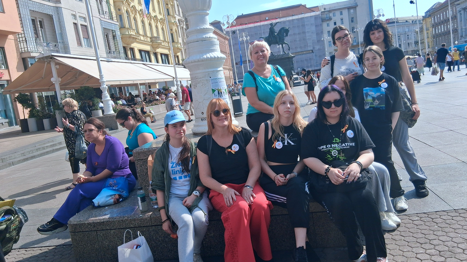 Grupa žena i djevojaka sjedi i stoji oko kamenog postolja na Trgu bana Jelačića u Zagrebu. Na grudima nose žute vrpce i bedževe kao znak sudjelovanja u Međunarodnoj akciji hodanja 45.000 koraka. U pozadini se vide prolaznici, kafići i kip bana Jelačića na konju.