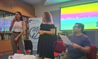 Tri osobe sudjeluju na događanju povodom Međunarodnog tjedna gluhih osoba i Međunarodnog dana znakovnog jezika. Žena lijevo prevodi na znakovni jezik, žena u sredini obraća se publici, a muškarac desno sjedi s papirom u ruci. Iza njih je projekcija s natpisom „Taj divni, šareni grad“ u duginim bojama te banner Hrvatskog saveza gluhih i nagluhih.