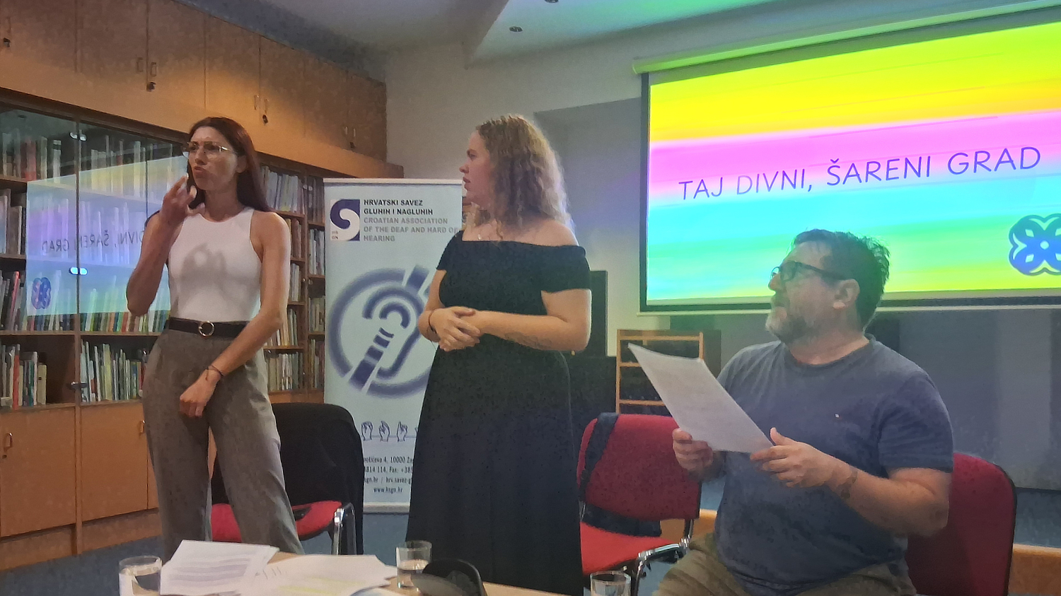 Tri osobe sudjeluju na događanju povodom Međunarodnog tjedna gluhih osoba i Međunarodnog dana znakovnog jezika. Žena lijevo prevodi na znakovni jezik, žena u sredini obraća se publici, a muškarac desno sjedi s papirom u ruci. Iza njih je projekcija s natpisom „Taj divni, šareni grad“ u duginim bojama te banner Hrvatskog saveza gluhih i nagluhih.