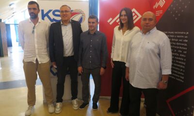 Fotografija prikazuije nekoliko ljudi lijepo obučenih u nekom prostoru. Iza njih stoje neki plakati.