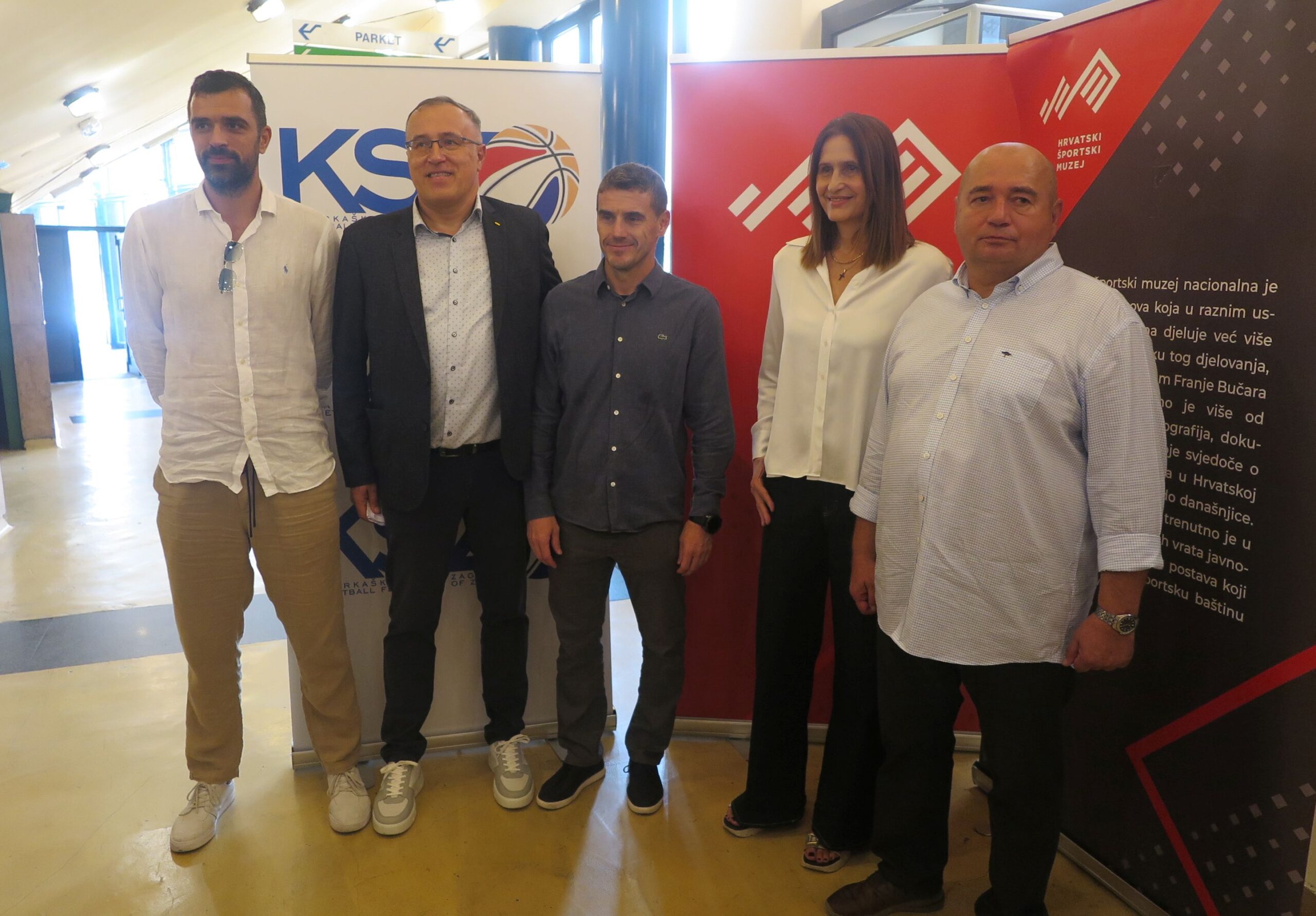 Fotografija prikazuije nekoliko ljudi lijepo obučenih u nekom prostoru. Iza njih stoje neki plakati.