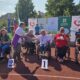 Fotografija prikazuje nekoliko ljudi u kolicima koji se nalaze na atletskoj stazi. Ispred njih nalaze se brojevi koji označavaju na kojem je mjestu tko završio. Neki od njih drže medalje u rukama.