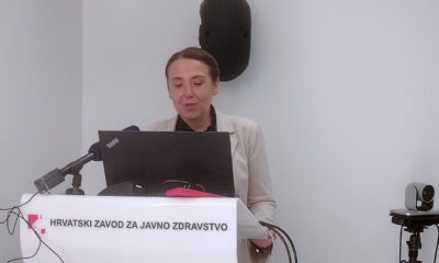 Žena u svijetloj jakni stoji za govornicom Hrvatskog zavoda za javno zdravstvo i drži izlaganje uz prijenos uživo. Na govornici piše 'Hrvatski zavod za javno zdravstvo'. Ispred nje je mikrofon i prijenosna kamera, a na zidu iza nje vise tehnička oprema i projekcijsko platno.
