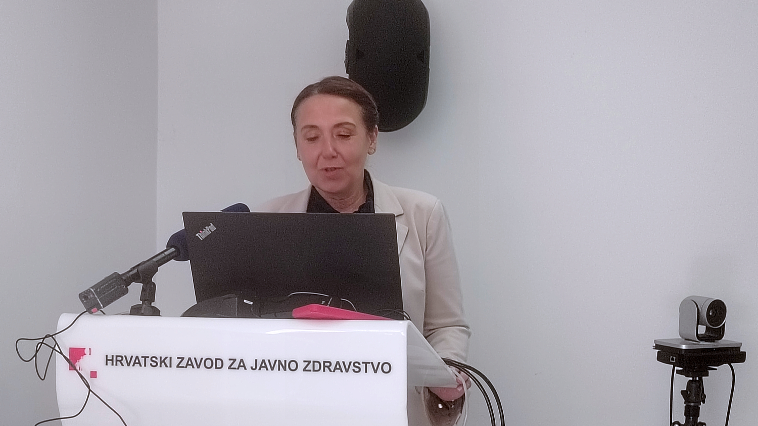 Žena u svijetloj jakni stoji za govornicom Hrvatskog zavoda za javno zdravstvo i drži izlaganje uz prijenos uživo. Na govornici piše 'Hrvatski zavod za javno zdravstvo'. Ispred nje je mikrofon i prijenosna kamera, a na zidu iza nje vise tehnička oprema i projekcijsko platno.