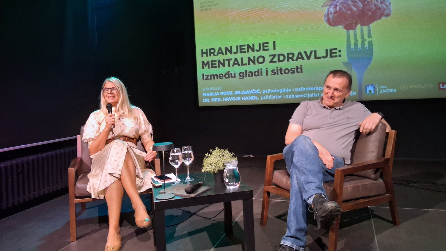 Psihologinja i psihoterapeutkinja Marija Roth Jelsaivić i psihijatar Hrvoje Handl sjede na pozornici tijekom okruglog stola o hranjenju i mentalnom zdravlju, s projekcijom naslova događanja u pozadini.