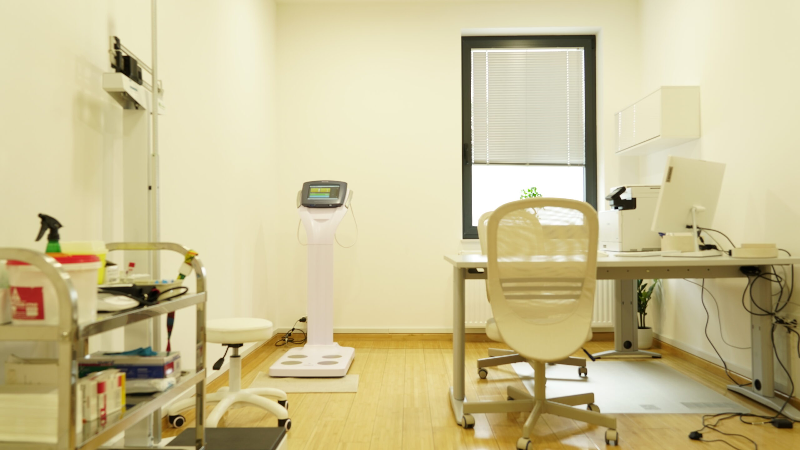 Na fotografiji je svijetla i uredna ordinacija modernog izgleda. U prostoru dominiraju svijetli tonovi zidova i namještaja, dok je pod od svijetlog drva. S lijeve strane nalazi se metalna kolica s medicinskim potrepštinama, poput rukavica, posuda, dezinfekcijskog sredstva i različitih pribora. U pozadini, nasuprot prozoru s roletama, vidi se uređaj za analizu sastava tijela s mjernom platformom i ekranom. Desno je radni stol s računalom, pisačem i uredskim priborom, ispred kojeg je moderna bijela stolica na kotačićima. Prostor djeluje profesionalno, minimalistički i higijenski, namijenjen pregledu i analizi zdravstvenog stanja pacijenata.