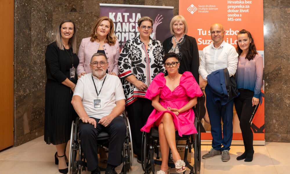 Skupna fotografija osmero ljudi snimljena u interijeru, ispred promotivnih panela Saveza društava multiple skleroze Hrvatske. Dvoje osoba u prvim redovima koriste invalidska kolica, jedna žena odjevena u bijelu košulju i muškarac u bijeloj majici. Uz njih stoji šestero osoba odjevenih u poslovno-ležernu odjeću. Svi se smiješe prema kameri.