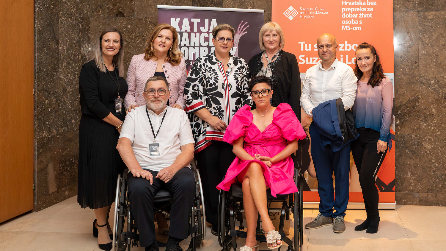 Skupna fotografija osmero ljudi snimljena u interijeru, ispred promotivnih panela Saveza društava multiple skleroze Hrvatske. Dvoje osoba u prvim redovima koriste invalidska kolica, jedna žena odjevena u bijelu košulju i muškarac u bijeloj majici. Uz njih stoji šestero osoba odjevenih u poslovno-ležernu odjeću. Svi se smiješe prema kameri.