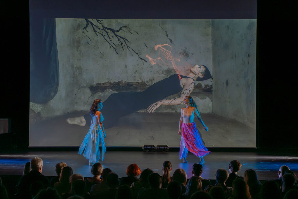 Scenska izvedba na pozornici s dvije plesačice u dugim, prozirnim suknjama u plavim i ljubičastim tonovima. U pozadini se projicira velika slika žene koja leži uz zid, s umjetničkim efektom dimnih linija koje izlaze iz njezina tijela. Publika sjedi u prvom planu, gledajući izvedbu.