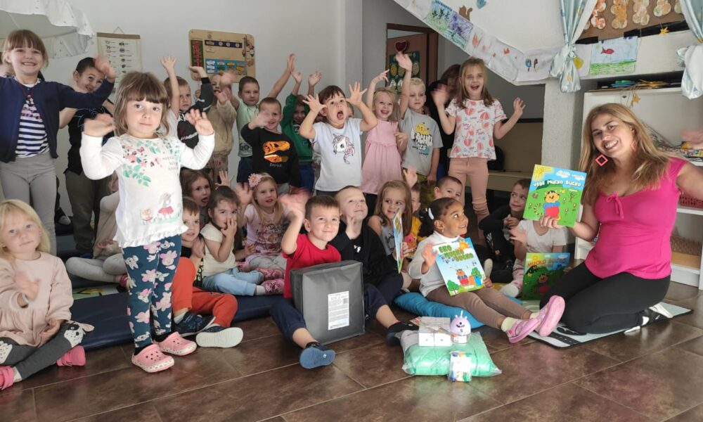 Fotografija prikazuje razigranu i srdačnu atmosferu u učionici, gdje se skupina male djece okupila za zajedničku fotografiju s odraslom ženom — vjerojatno učiteljicom ili odgajateljicom. Djeca su nasmijana, podignutih ruku, zračeći uzbuđenjem i povjerenjem. Žena sjedi na podu ispred njih, držeći dvije šarene dječje knjige, što dodatno naglašava edukativni kontekst i poticajnu energiju prostora. Na podu ispred grupe nalaze se razni predmeti — paketići, igračke i edukativni materijali — koji doprinose dojmu da se radi o posebnoj aktivnosti ili proslavi. Zidovi učionice ukrašeni su dječjim crtežima i edukativnim plakatima, stvarajući šarenu i poticajnu okolinu koja slavi kreativnost i zajedništvo.