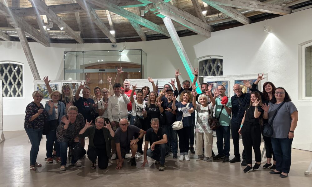 Na slici se nalazi velika skupina ljudi okupljena u svijetlom prostoru s visokim drvenim stropom i vidljivim gredama. Među okupljenima su muškarci i žene različitih dobi, mnogi od njih nasmijani i mašu prema kameri. Prostor podsjeća na galeriju ili muzej — zidovi su bijeli, pod je uglačan, a na zidovima vise uokvirene umjetnine. Veliki prozori s dekorativnim rešetkama propuštaju prirodnu svjetlost, stvarajući ugodnu atmosferu. Ljudi su raspoređeni u polukrugu: neki kleče ili sjede sprijeda, dok ostali stoje iza njih.