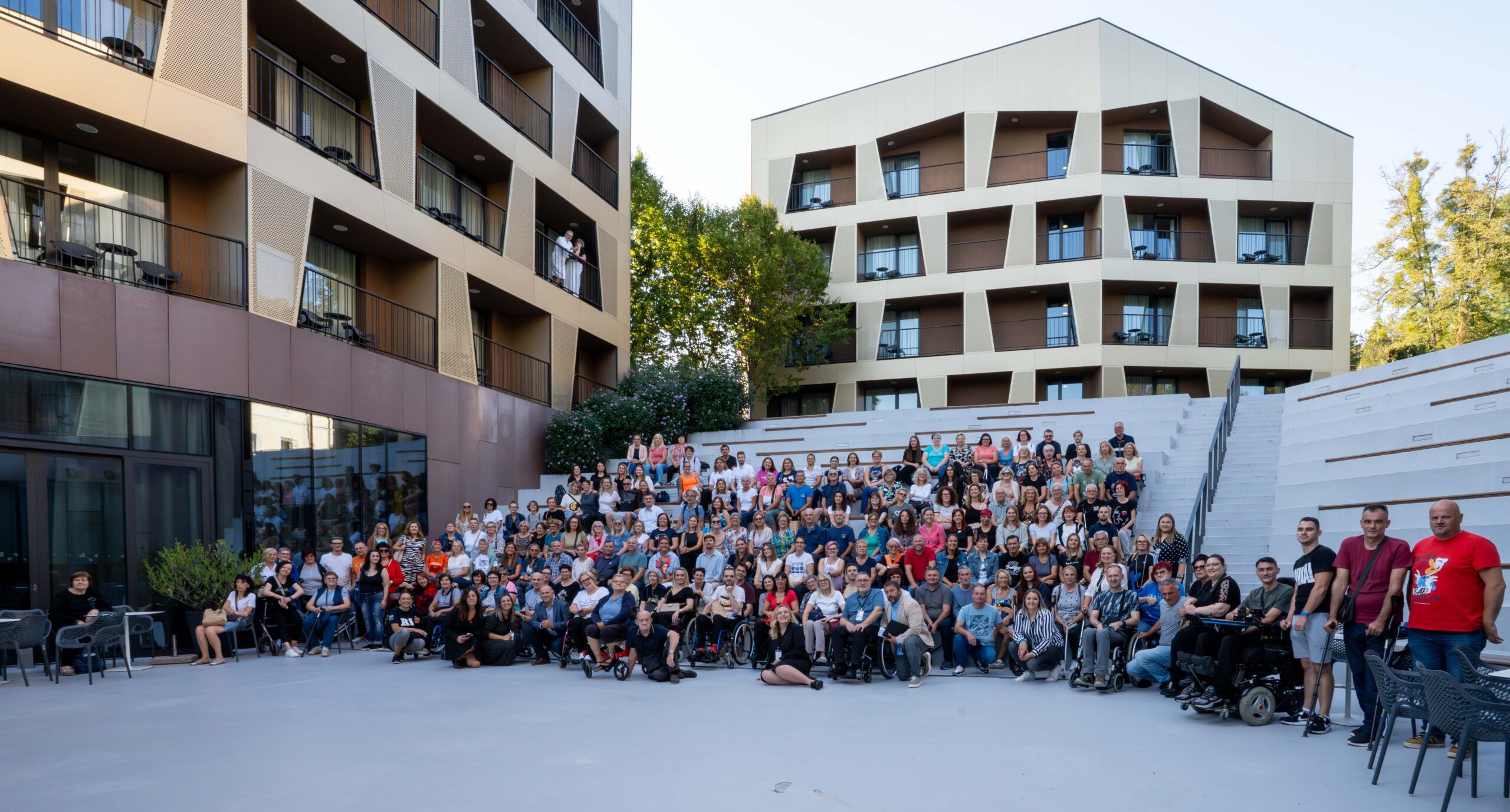 Na fotografiji je velika skupina ljudi okupljena ispred modernog hotelskog kompleksa s više katova i balkonima. Snimljeni su na vanjskom amfiteatru, raspoređeni u nekoliko redova na bijelim tribinama i na prostoru ispred njih. U prvom planu vide se sudionici u invalidskim kolicima, dok iza njih stoji i sjedi mnoštvo ljudi različite dobi. Atmosfera djeluje svečano i zajednički, kao uspomena na okupljanje ili događaj. U pozadini se vidi zelenilo, a s balkona hotela nekoliko osoba promatra prizor. Fotografija prenosi osjećaj zajedništva, podrške i pozitivne energije velike grupe ljudi.