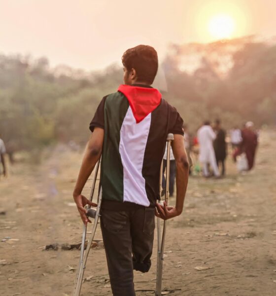 Fotografija prikazuje osobu bez noge koja hoda s obje štake. Na sebi ima kratku majicu, kratke hlače, a iza leđa crvenu maramu.