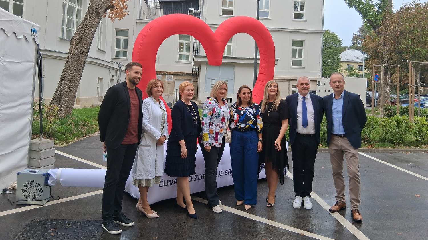 Skupina od osam ljudi pozira ispred velike crvene instalacije u obliku srca na otvorenom prostoru, ispred bijele zgrade. Riječ je o sudionicima događaja posvećenog očuvanju zdravlja srca.