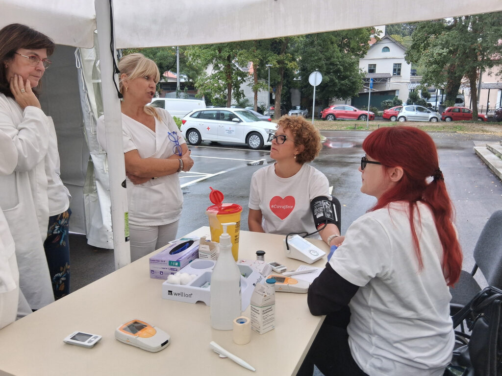 Dva medicinska djelatnika u bijelim kutama razgovaraju s dvjema volonterkama koje nose bijele majice s crvenim srcem i natpisom „#Čuvajsrce“. Jedna volonterka mjeri krvni tlak drugoj osobi, a na stolu ispred njih nalaze se medicinski uređaji i potrošni materijal.