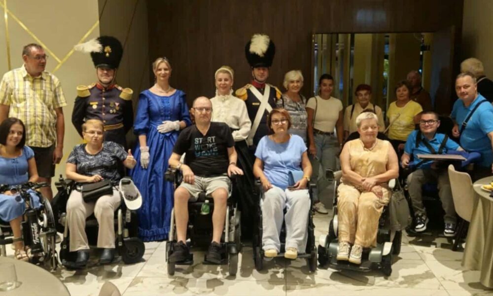 Na fotografiji se nalazi skupina od 18 osoba okupljenih u zatvorenom prostoru, vjerojatno tijekom društvenog ili zajedničkog događanja. Među njima su i osobe u invalidskim kolicima, što ukazuje na inkluzivnost i otvorenost zajednice. Posebnu pažnju privlače dvije osobe odjevene u povijesne vojne uniforme te jedna žena u plavoj povijesnoj haljini, što sugerira da se radi o tematskom događaju ili povijesnoj rekonstrukciji.