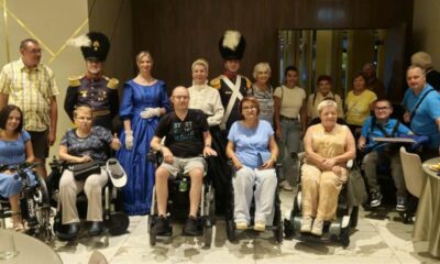 Na fotografiji se nalazi skupina od 18 osoba okupljenih u zatvorenom prostoru, vjerojatno tijekom društvenog ili zajedničkog događanja. Među njima su i osobe u invalidskim kolicima, što ukazuje na inkluzivnost i otvorenost zajednice. Posebnu pažnju privlače dvije osobe odjevene u povijesne vojne uniforme te jedna žena u plavoj povijesnoj haljini, što sugerira da se radi o tematskom događaju ili povijesnoj rekonstrukciji.