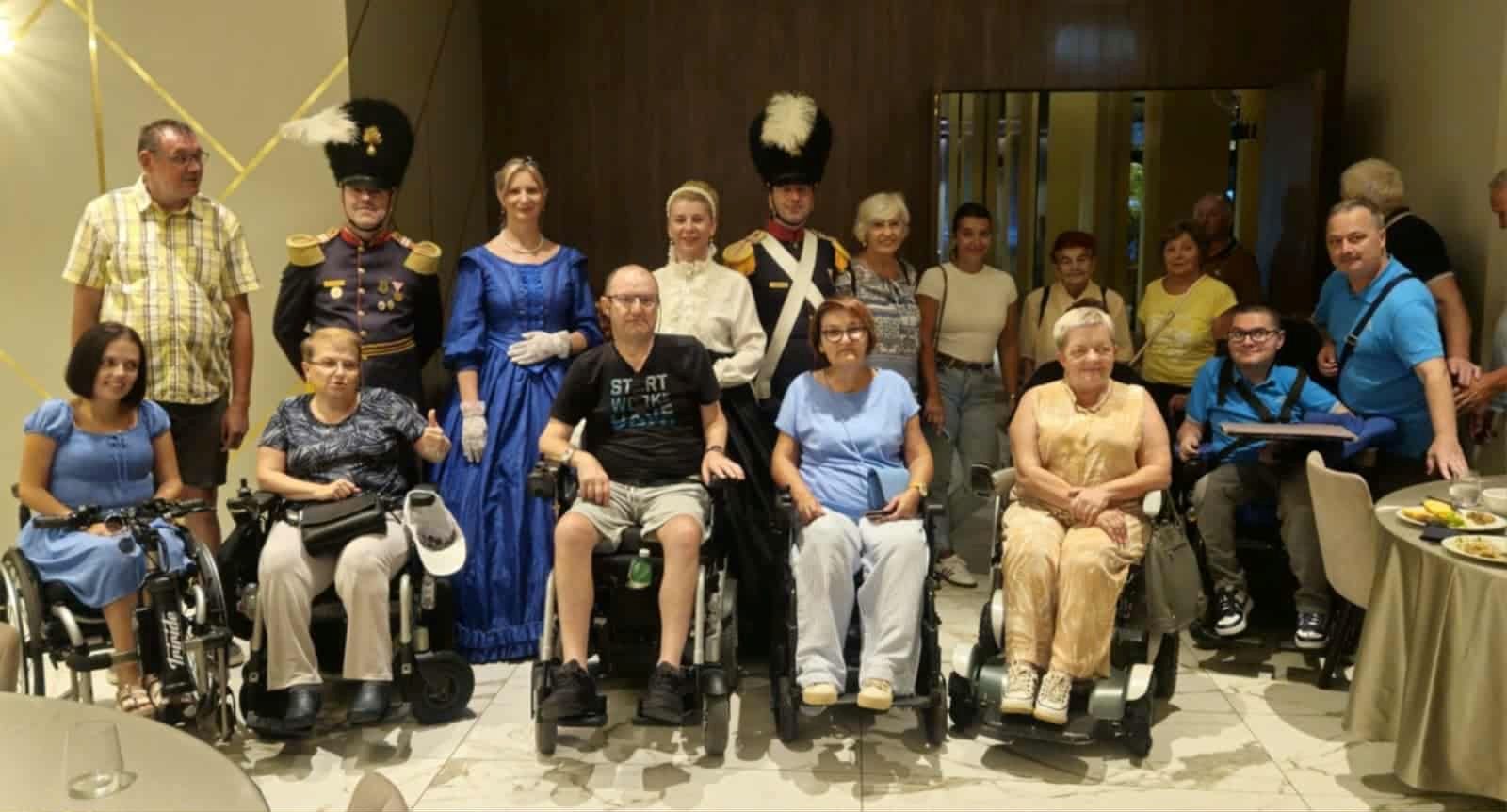 Na fotografiji se nalazi skupina od 18 osoba okupljenih u zatvorenom prostoru, vjerojatno tijekom društvenog ili zajedničkog događanja. Među njima su i osobe u invalidskim kolicima, što ukazuje na inkluzivnost i otvorenost zajednice. Posebnu pažnju privlače dvije osobe odjevene u povijesne vojne uniforme te jedna žena u plavoj povijesnoj haljini, što sugerira da se radi o tematskom događaju ili povijesnoj rekonstrukciji.