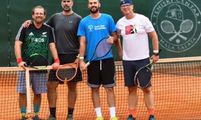 Fotografija prikazuje tenisače kako stoje na pješčanom teniskom terenu. Svi u ruci imaju rekete.