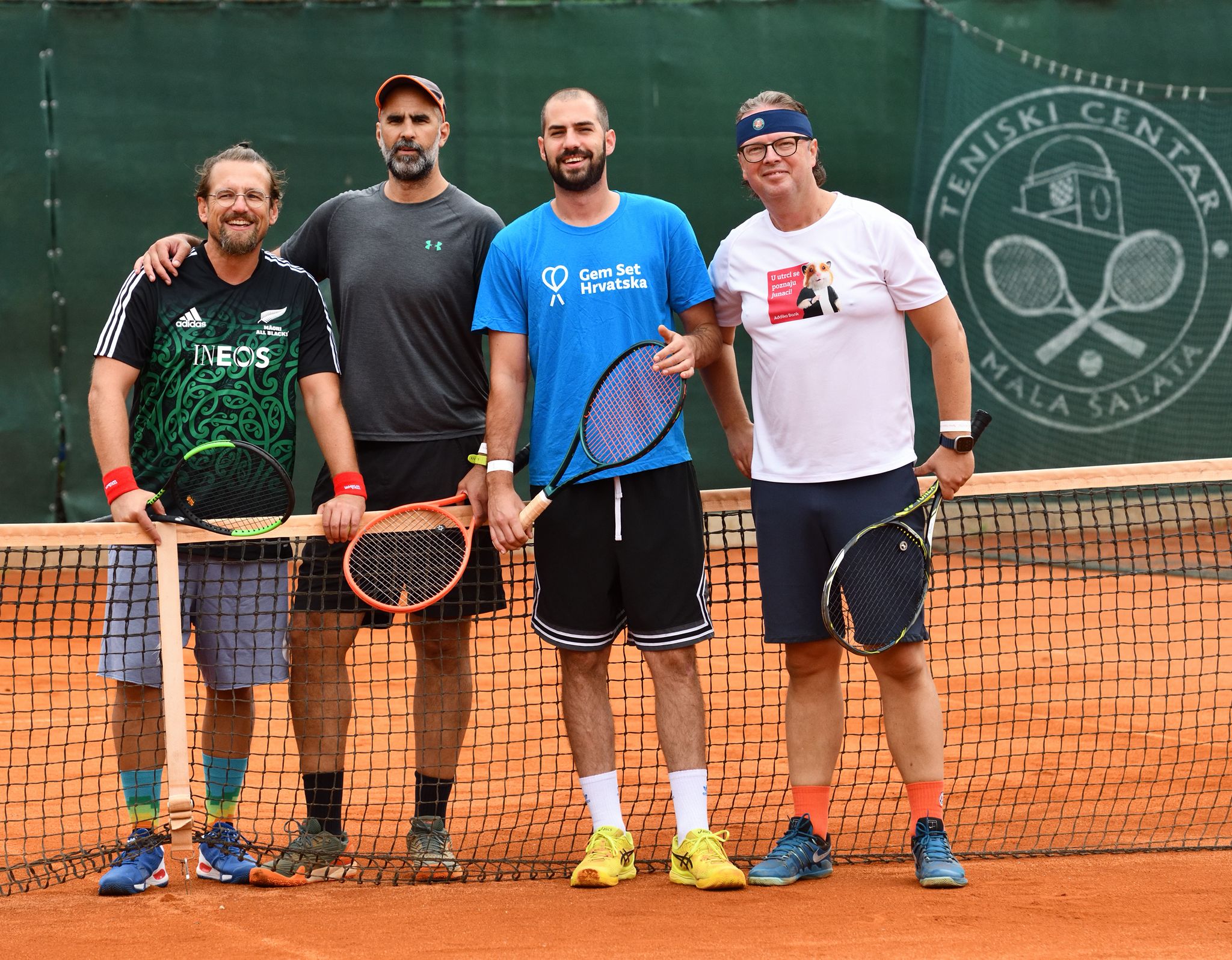 Fotografija prikazuje tenisače kako stoje na pješčanom teniskom terenu. Svi u ruci imaju rekete.