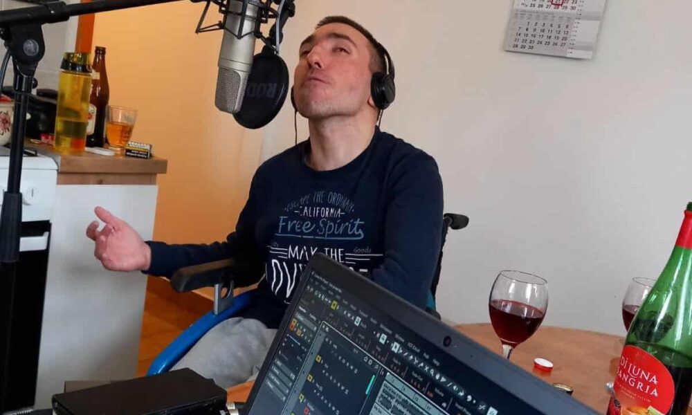 Na fotografiji je muškarac u invalidskim kolicima koji sjedi ispred mikrofona i nosi slušalice. Djeluje kao da je u studijskom okruženju, usred snimanja ili probe – ispred njega je otvoreno računalo s glazbenim programom, a u pozadini se vidi kuhinjski prostor. Na stolu se nalaze čaše s crnim vinom i boca sangrije, što daje opuštenu i prijateljsku atmosferu. Njegov izraz lica i položaj ruku odaju dojam da je potpuno predan glazbi i trenutku, kao da pjeva s puno emocija i užitka.