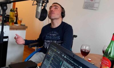 Na fotografiji je muškarac u invalidskim kolicima koji sjedi ispred mikrofona i nosi slušalice. Djeluje kao da je u studijskom okruženju, usred snimanja ili probe – ispred njega je otvoreno računalo s glazbenim programom, a u pozadini se vidi kuhinjski prostor. Na stolu se nalaze čaše s crnim vinom i boca sangrije, što daje opuštenu i prijateljsku atmosferu. Njegov izraz lica i položaj ruku odaju dojam da je potpuno predan glazbi i trenutku, kao da pjeva s puno emocija i užitka.