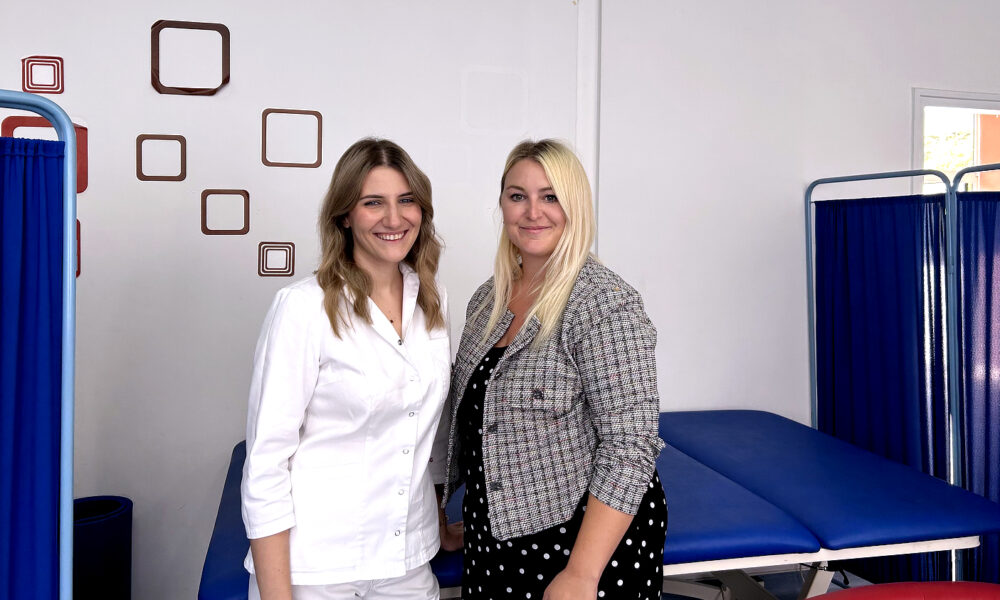 Dvije žene stoje nasmiješene u prostoriji za fizikalnu terapiju. Lijevo je žena u bijeloj medicinskoj odori, a desno žena u crnoj haljini s bijelim točkicama i kariranom jaknom. Iza njih se nalaze plavi terapeutski ležajevi i paravani.